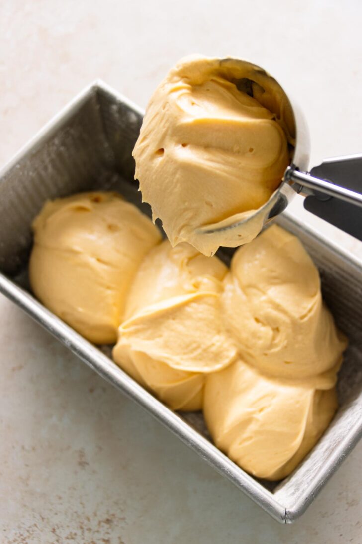 Using an ice cream scoop to fill the prepared loaf pan with pound cake batter.