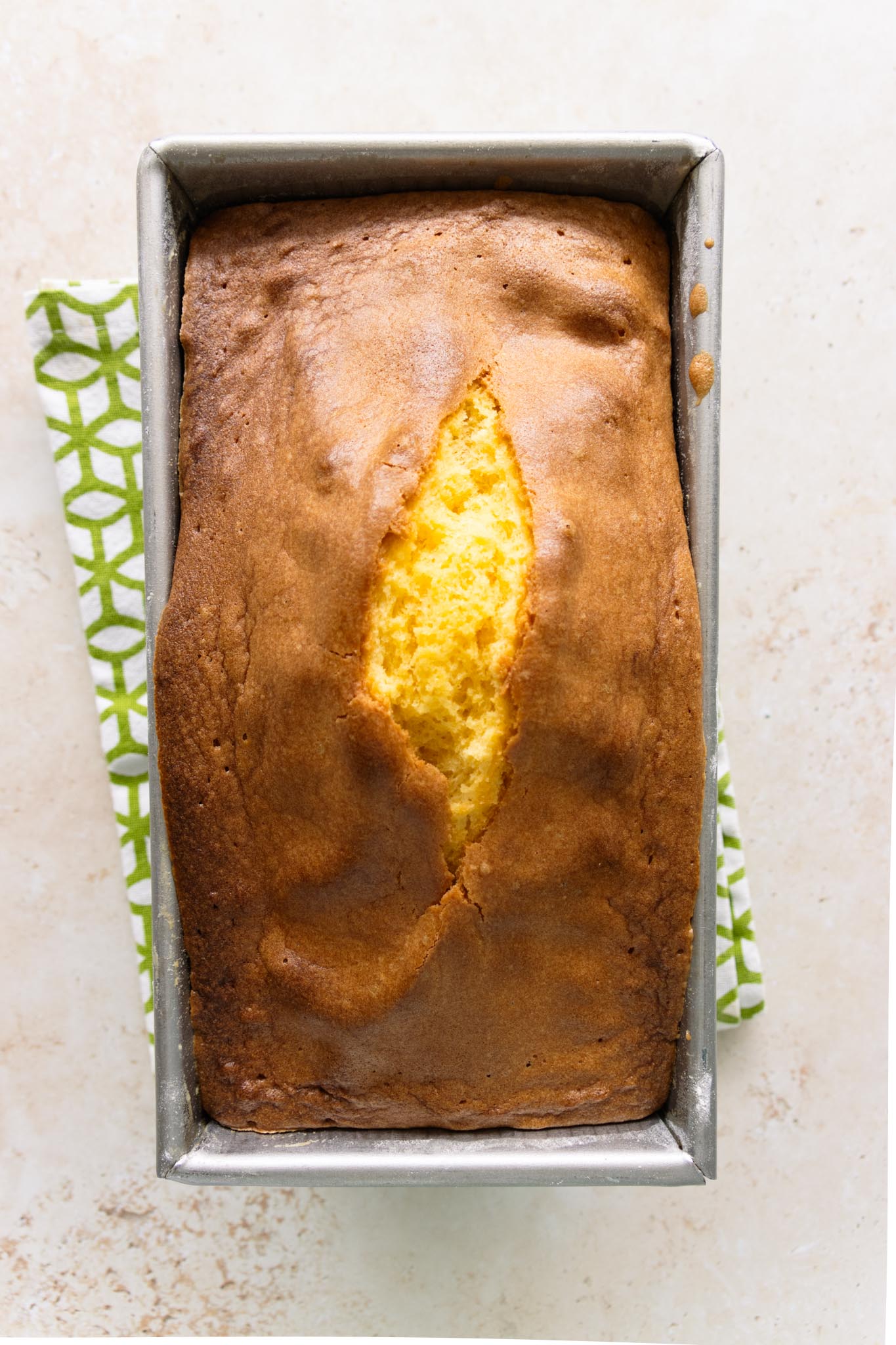 The baked pound cake baked to a golden brown crust, split on the top showing the yellow buttery crumb, cooling in the loaf pan set upon a tea towel.