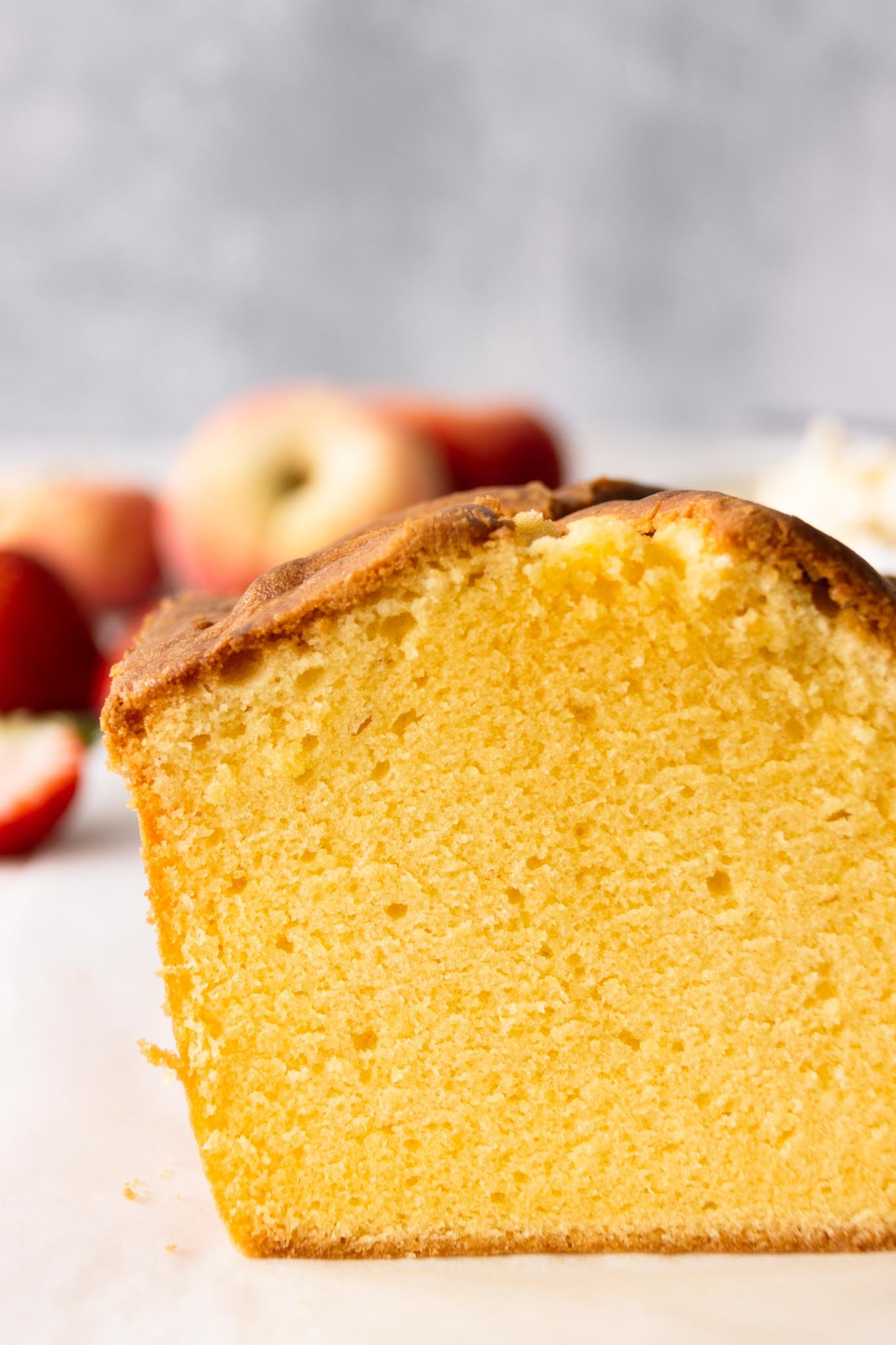 Close-up view of a pound cake slice, showing the moist, buttery, and velvety smooth crumb with fresh strawberries and peaches alongside a bowl of whipped cream in the background.