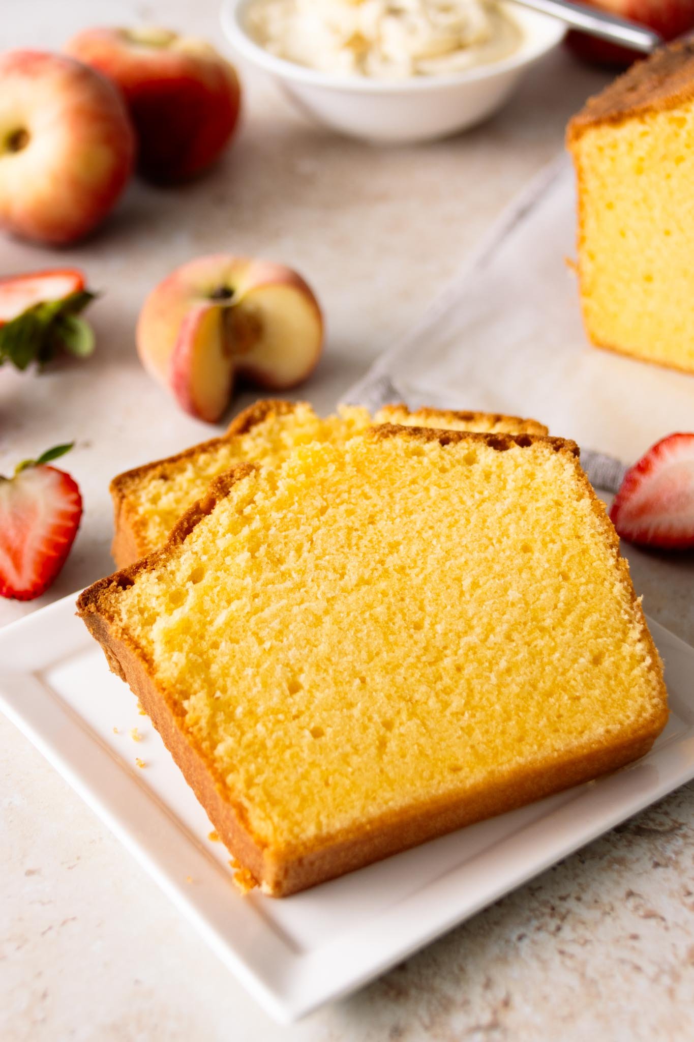 Two pound cake slices set on a sqaure serving plate, showing the moist, buttery, and velvety smooth crumb with fresh strawberries and peaches alongside a bowl of whipped cream in the background.