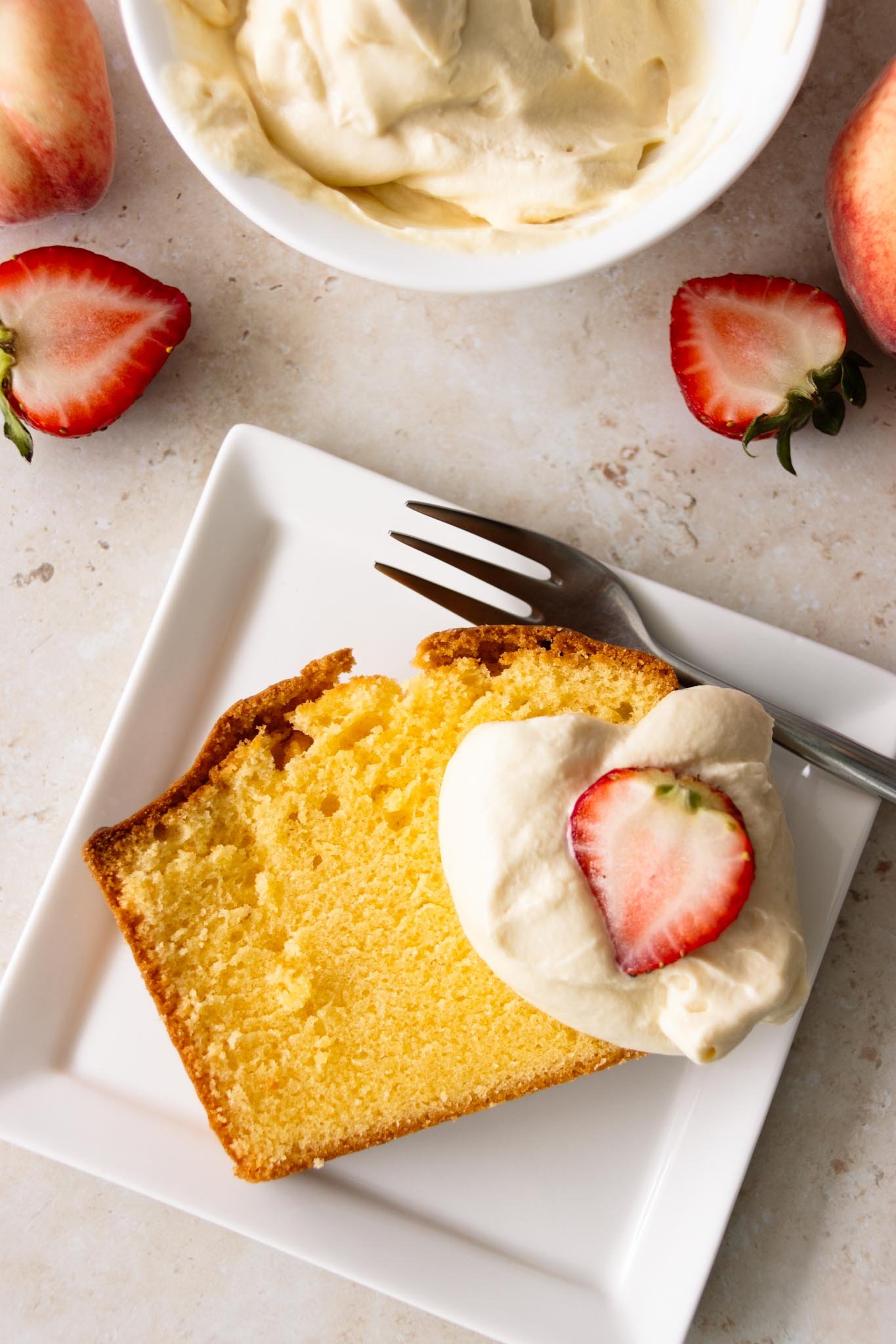 A pound cake slice topped with a dollop of whipped cream and fresh strawberry halves, set on a square serving plate with a dessert fork.